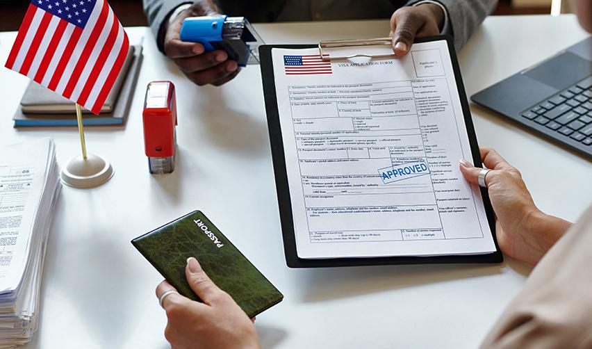 A person holding a U.S. application document for green cards, with a stamp and an American flag on a desk and a passport in her hand.