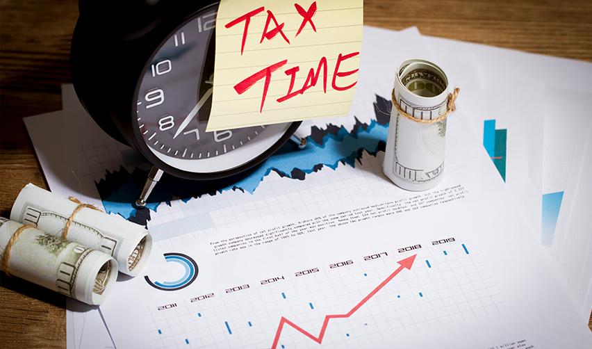 Some new commercial enterprise documents and graphs on a desk, alongside business investment money, a clock, and a note saying tax time.