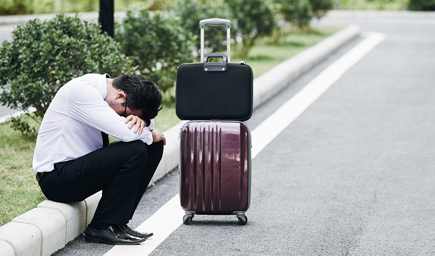 A person sitting on a curb with his head down, with luggage on his side.