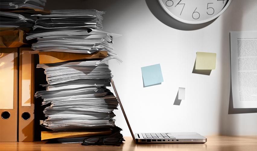 A computer and a big pile of papers on a desk at an office.