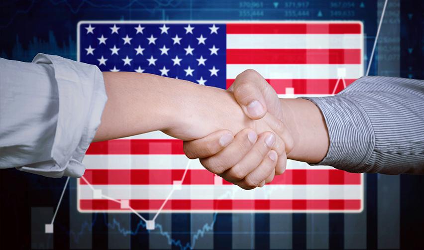 A close-up of hands shaking in front of a U.S. flag and some business graphs.