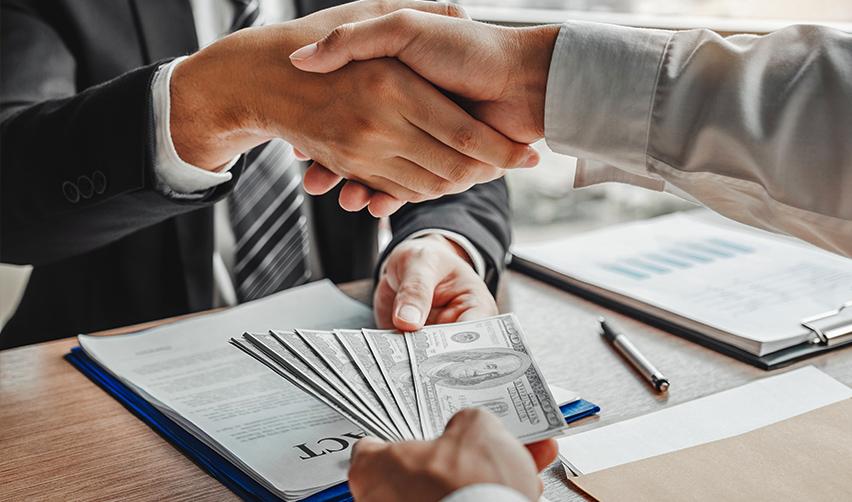 Two hands shaking and exchanging some money above a contract on a desk.
