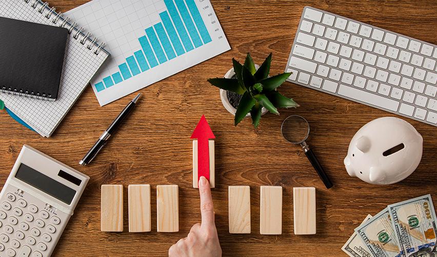 A person pushing a wooden block up on a desk with money and business items on it, symbolizing the benefits of EB5 for developers.