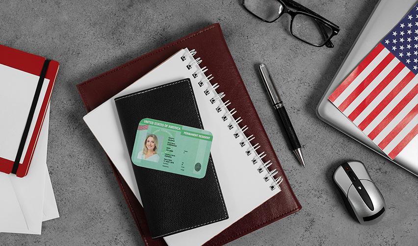 Notebooks, a computer and mouse, and a pen on a desk, with a U.S. Permanent Resident Card (green card) on the top.