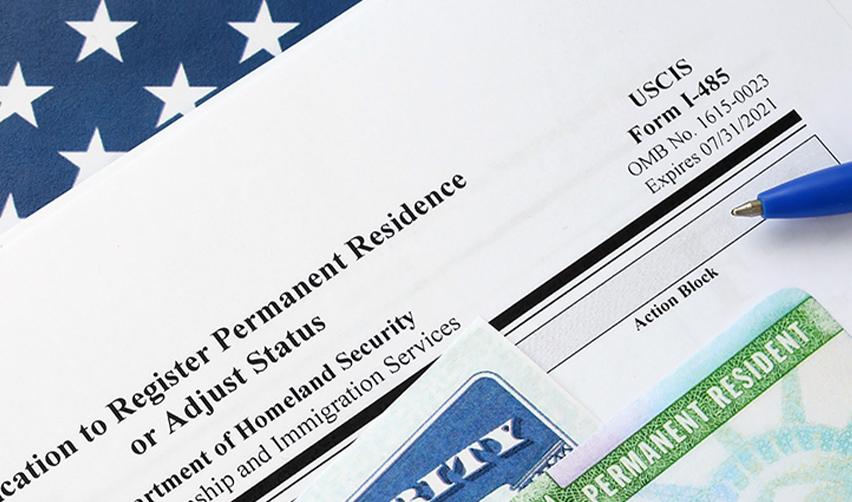 Close-up of a Form I-485, Application to Register Permanent Residence or Adjust Status, on a desk under a Green Card.