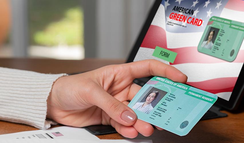 A person holding a United States of America Permanent Resident card in her hand in front of a computer, symbolizing employment based adjustment of status.