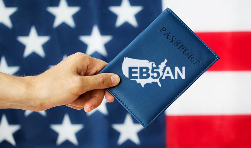 A hand holding a blue passport with the EB5AN logo on it in front of the American flag.