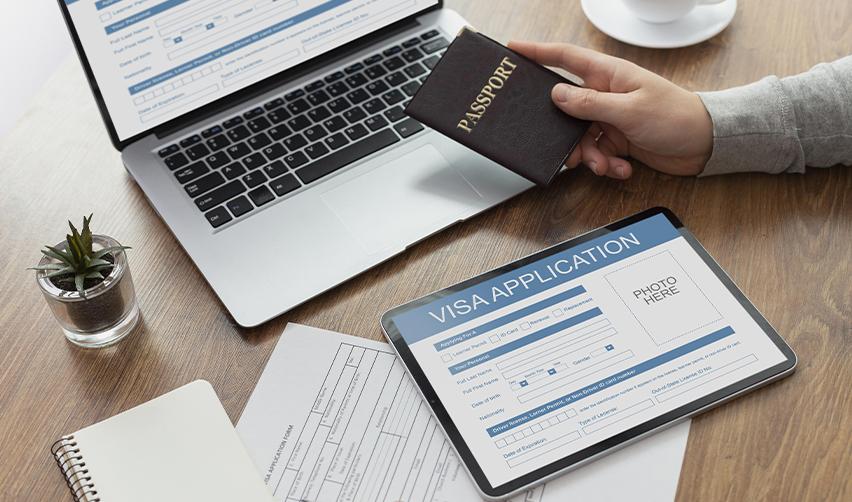 A hand holding a passport above some visa application documents and forms.