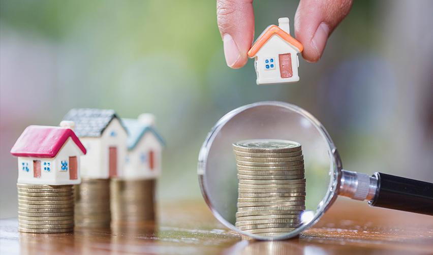 A hand holding a small house over a stack of coins behind a magnifying glass. EB5AN can help foreign investors who want to use HELOCs as their EB5 source of funds.