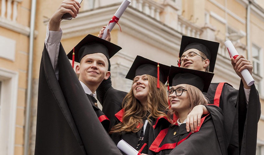 A group of college students taking a selfie in front of their university in graduation caps and robes.