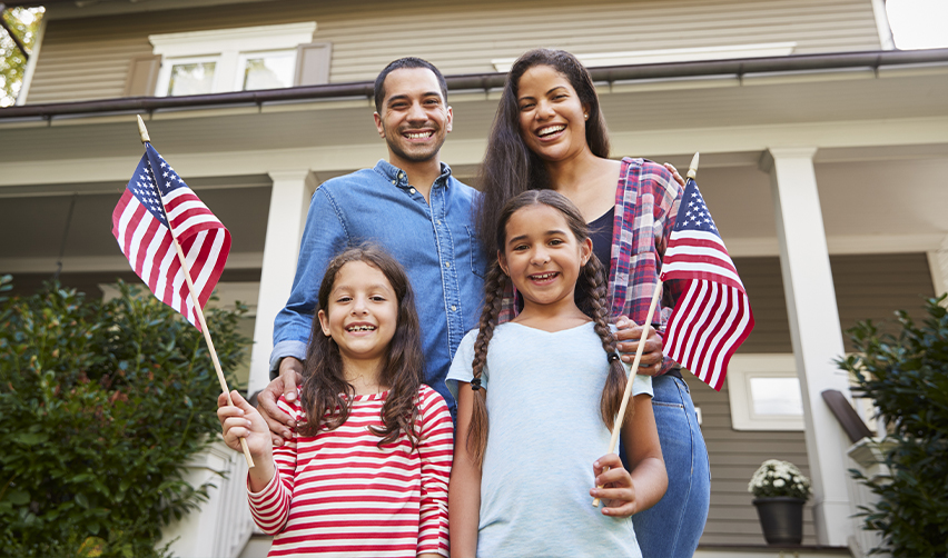 Smiling family with two children holding American flags in front of a home, representing the EB5 green card pathway to permanent residency and achieving the American Dream.