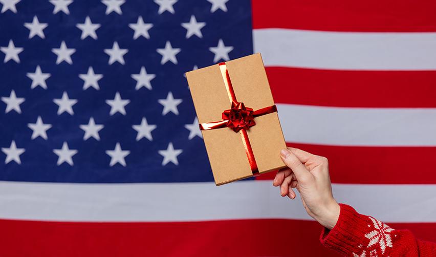 A hand holding a gift-wrapped box with a red ribbon in front of an American flag, symbolizing the gift of a US Green Card through the EB5 program with the assistance of EB5AN.