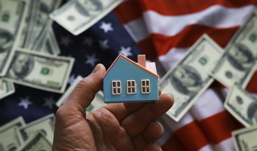 A hand holding a small house model with US dollar bills and an American flag in the background, symbolizing investment opportunities in the US economy through the EB5 program.