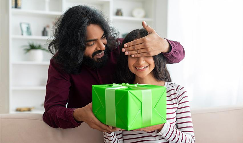 A father surprising his daughter with a green gift box, representing the opportunity to secure a brighter future for families through the EB5 Green Card program.