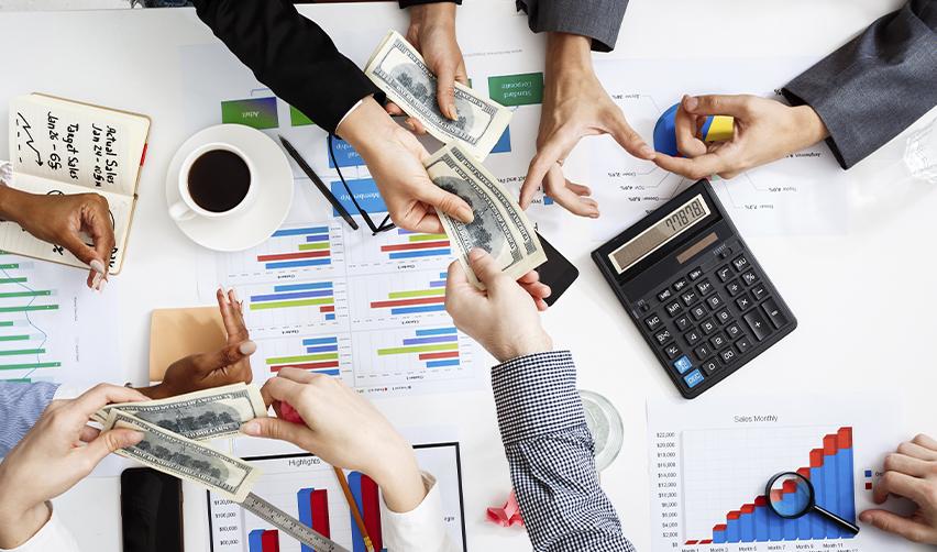 Multiple hands exchanging money over a desk filled with charts, graphs, and financial documents, representing portfolio diversification and investment planning in the EB5 program.