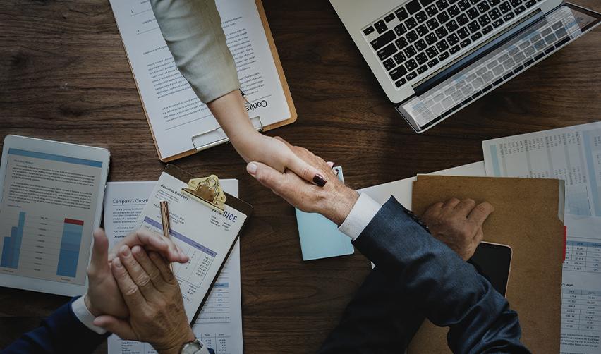 Two individuals shaking hands over a desk with documents and financial reports. Formal agreement and partnership for EB5 investment, emphasizing the importance of collaboration with immigration attorneys and regional centers.