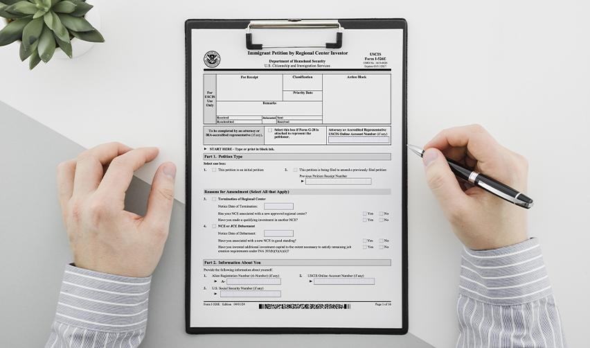 Hands filling out Form I-526E on a clipboard, showcasing the paperwork required for filing an EB5 immigrant petition.