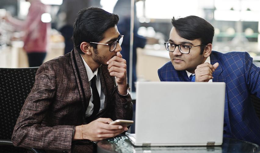 Two professionally dressed businessmen discussing visa options at a café, with a laptop open. Comparing H1B job sponsorship challenges with the secure pathway of the EB5 investor visa.