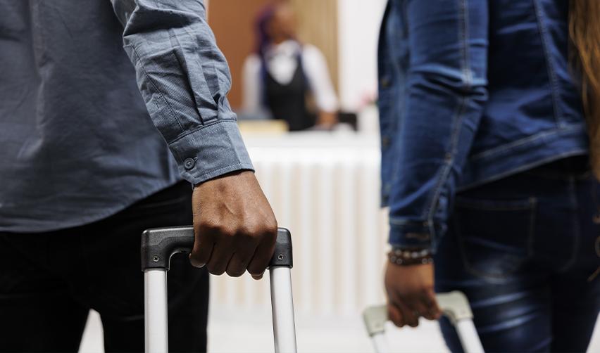 Two individuals holding suitcases stand at a reception desk, possibly checking into an immigration checkpoint. The changing immigration policy under the Trump administration may affect the ease of travel and visa processes for non-citizens.