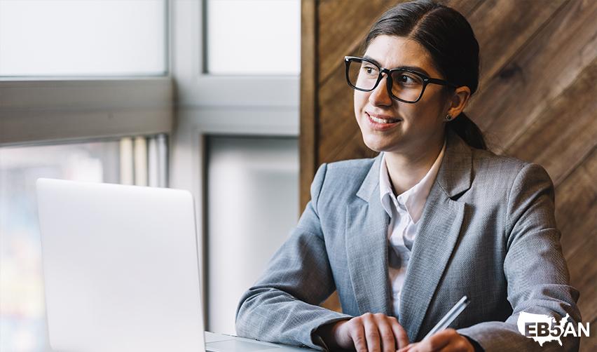 The EB5AN logo next to an international student in business attire, smiling while working on a laptop, representing the transition from an F1 visa holder to a U.S. Green Card holder through the EB5 program.