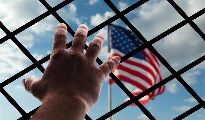 A hand reaches through a metal fence towards the American flag, symbolizing challenges in immigration enforcement and border control under new policies.