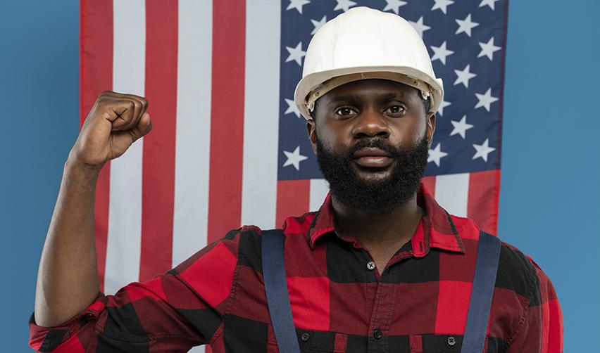 A determined construction worker wearing a white hard hat, standing in front of an American flag with his fist raised. The EB5 Immigrant Investor Program helps fund infrastructure projects, creating jobs in construction and skilled labor industries across the U.S.