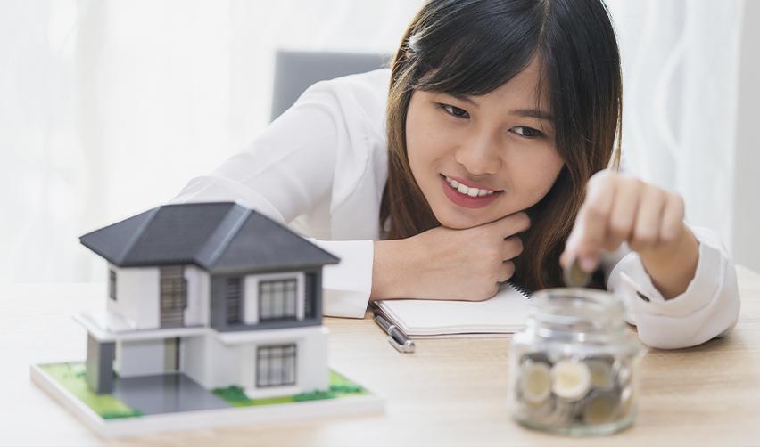 A young woman smiles while placing coins into a jar, with a model house on the table, symbolizing the use of home equity loans (HELOC) to fund EB5 investments.