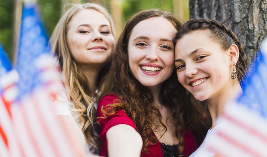 A group of young women smiling and waving American flags outdoors. The EB5 program not only provides foreign investors with a path to U.S. residency but also strengthens local communities by driving economic growth and job creation.