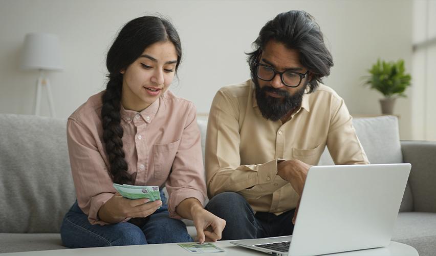 Indian couple reviewing documents and finances on a laptop, ensuring they check their approval notice on the United States Citizenship and Immigration Services website in a timely manner.