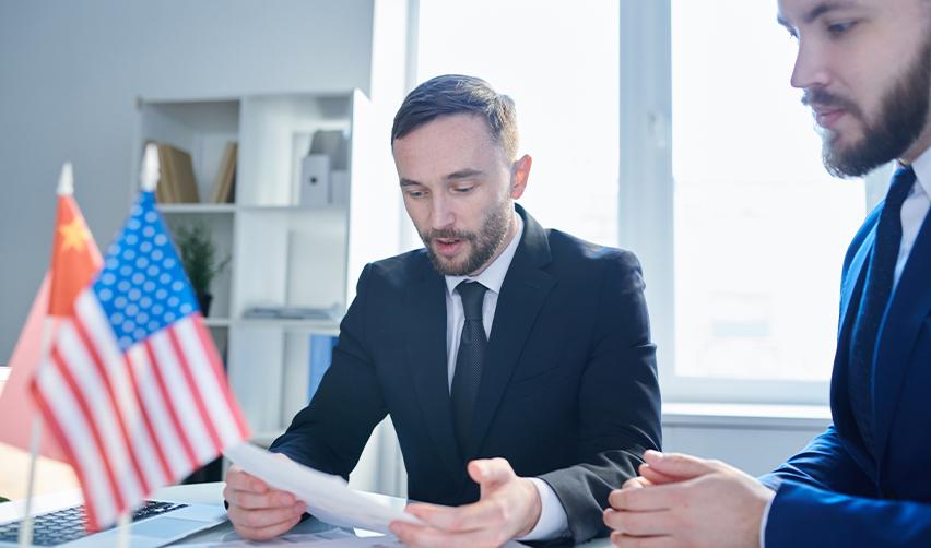 Businessmen in a visa consultation discussing EB5 new commercial enterprise requirements under U.S. immigration law, with international flags in the background.