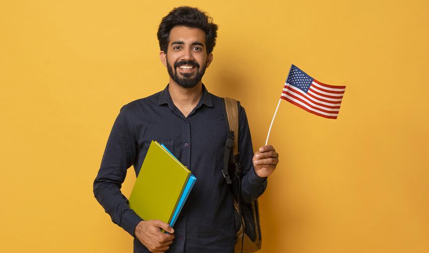 A smiling Indian student holding books and a small U.S. flag, representing educational opportunities available through the EB5 visa and US permanent residency.