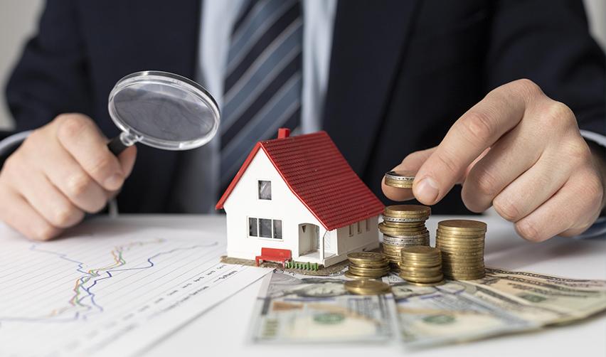 Close-up of a miniature house with stacked coins, dollar bills, and a magnifying glass, emphasizing capital investment and return potential in EB5AN real estate projects.