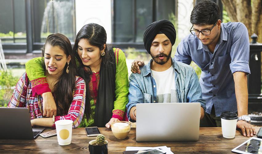 Group of young Indian professionals collaborating on laptops in an outdoor setting, discussing immigration options through a sponsoring employer that USCIS approves to obtain permanent residence.