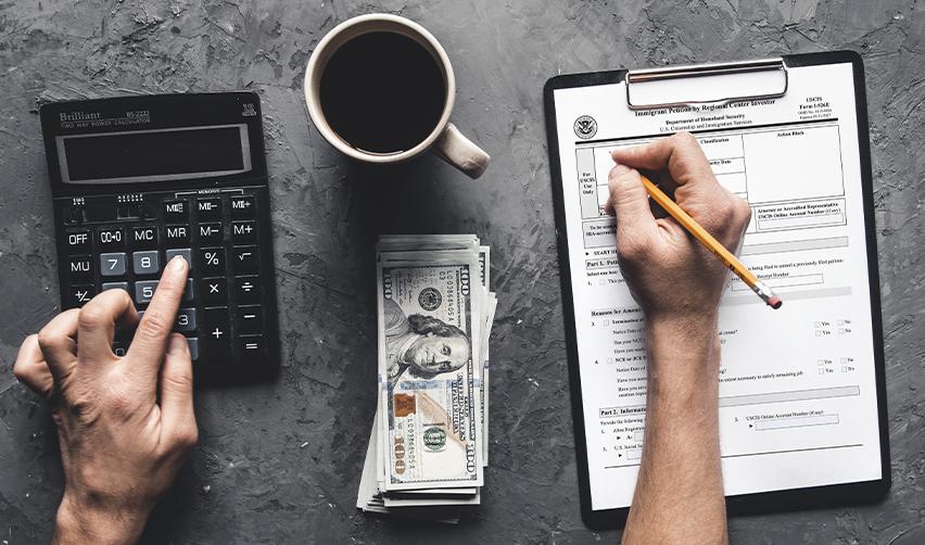 Hands using a calculator next to USCIS EB5 investment forms and stacks of hundred-dollar bills, highlighting financial planning and visa filing costs for EB5 investors.