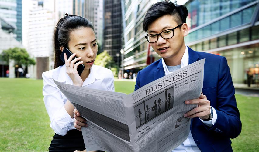 Asian EB5 investors discussing global investment news and visa opportunities while reviewing a business newspaper, highlighting rising interest in rural projects.