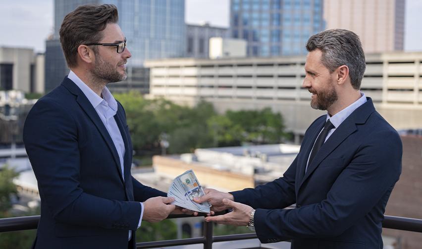 Two businessmen in suits exchanging a bundle of U.S. dollar bills, illustrating capital transfer and fund release in EB5 investment transactions.