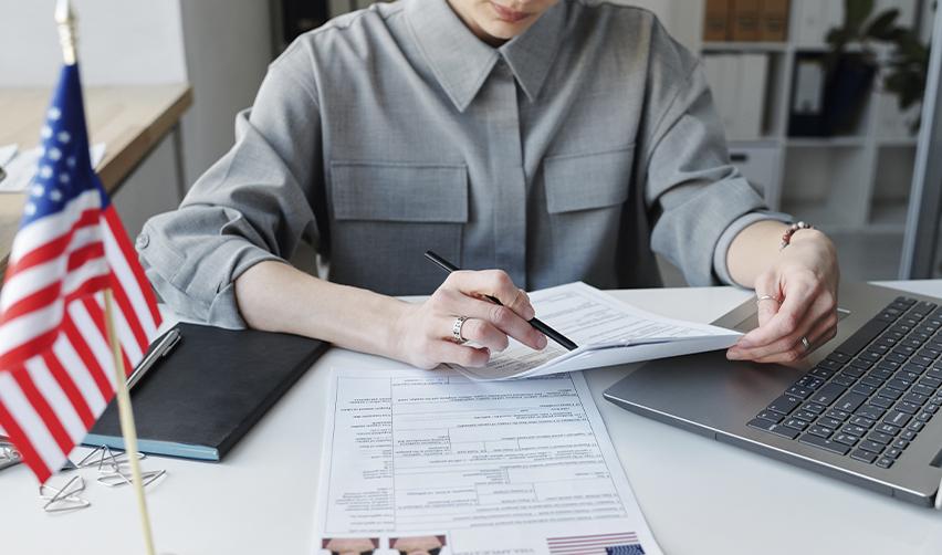 Woman filling out immigration paperwork with a laptop and U.S. flag on desk, representing an EB5 investor preparing forms like I-485 for adjustment of status.