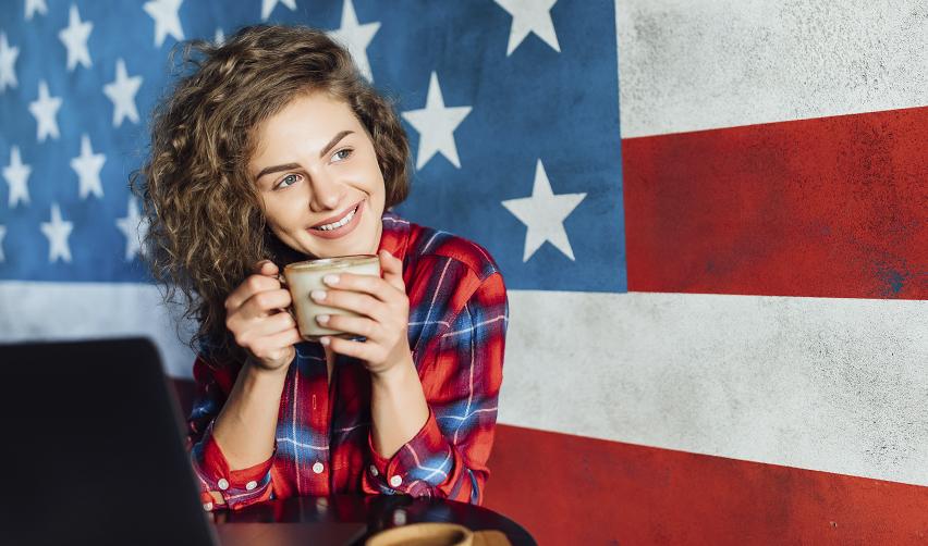 A smiling young woman in front of a large American flag mural, representing the freedom and comfort of living in the U.S. while awaiting EB5 Green Card approval.