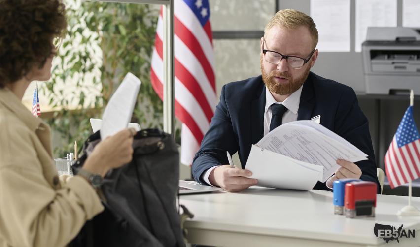 USCIS officer reviewing documents during an immigration interview, with American flags in view, illustrating consular processing for EB5 visa applicants.