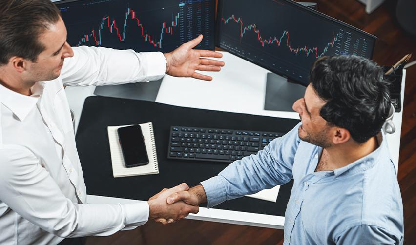 Business partners shaking hands in front of financial monitors, reflecting investor trust and collaboration in EB5 regional center job-creating entity projects.