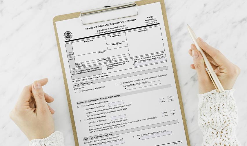 Hands holding a completed USCIS Form I-526E on a clipboard, representing the EB5 petition process for regional center investors in a rural new commercial enterprise.