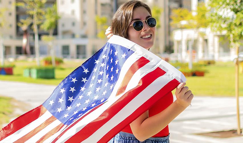 A smiling woman holding a U.S. flag in a public space, representing foreign nationals visiting the United States and their connection to American culture.