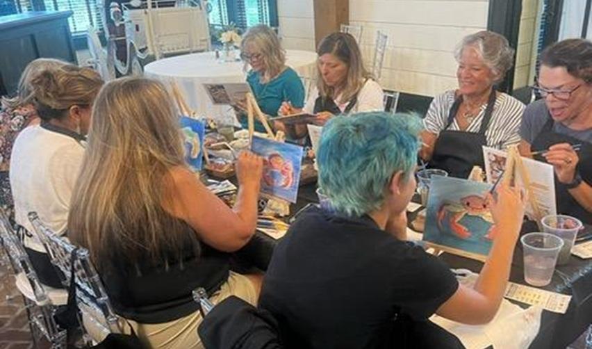 Group of Bay Creek residents smiling while painting coastal scenes at a guided art class, part of the community’s creative lifestyle programs.