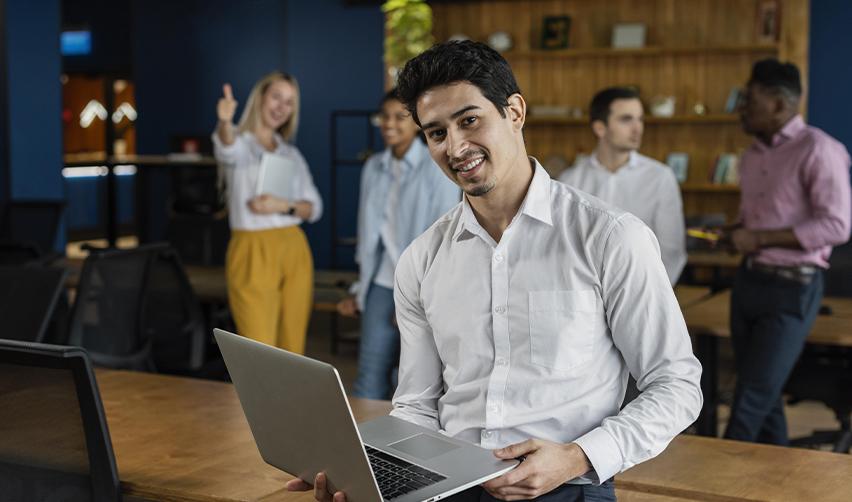 Smiling professional holding a laptop in a modern office setting with colleagues in the background, symbolizing career independence and stability through the EB5 program.