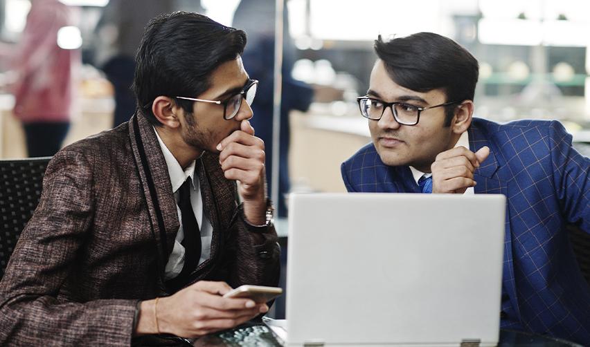 Two Indian professionals in suits discussing while looking at a laptop, representing career uncertainty and immigration planning for H1B employees.