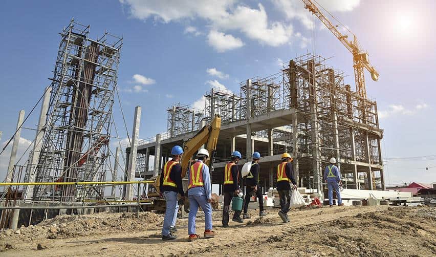 Construction workers in safety vests and helmets walking at an active building site with cranes and scaffolding, illustrating EB5 projects under development that create U.S. jobs.