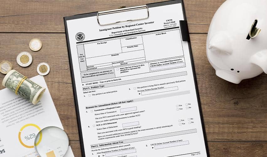 EB5 Form I526E on a clipboard beside coins, dollar bills, and a piggy bank, representing EB5 petition filing and investment documentation.