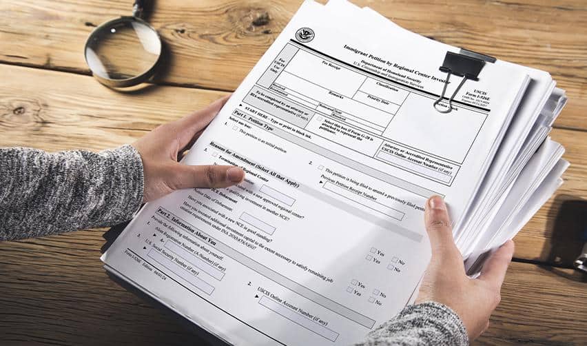 Hands holding a stack of USCIS Form I-526E petitions on a wooden desk beside a magnifying glass, representing EB5 application paperwork and filing process.