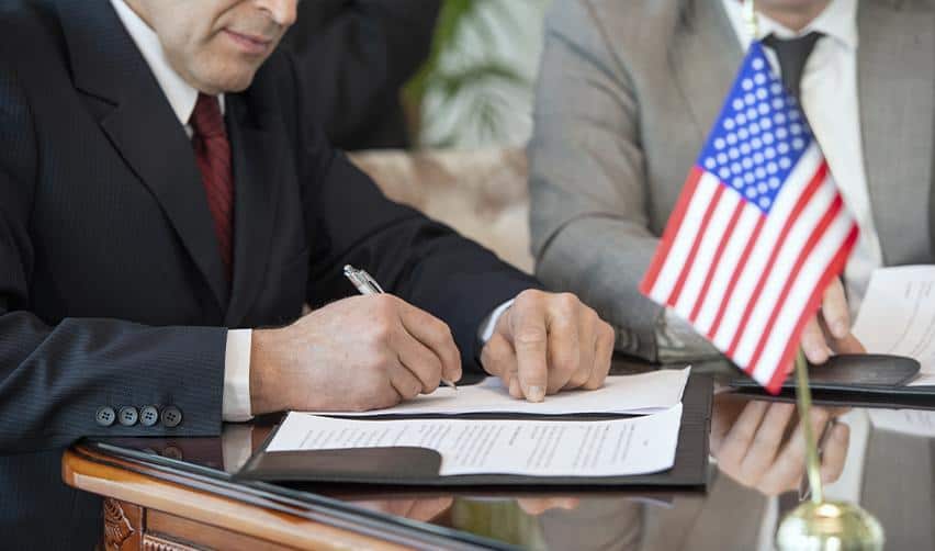 Two professionals in suits signing documents beside a small American flag, symbolizing USCIS approval and compliance for the Grand Park rural EB5 project.
